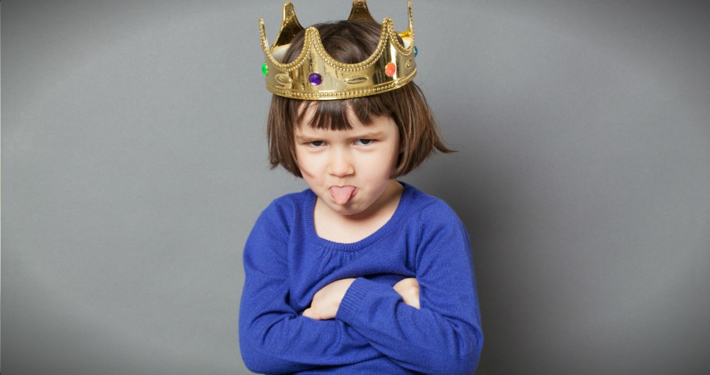 Child wearing a crown with her arms folded and sticking out her tongue